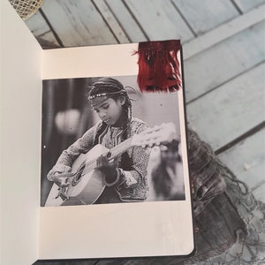 May include: A black and white photo of a person playing a guitar. The person is wearing a beaded headband and a patterned shirt. The photo is in a scrapbook with a red feather bookmark.