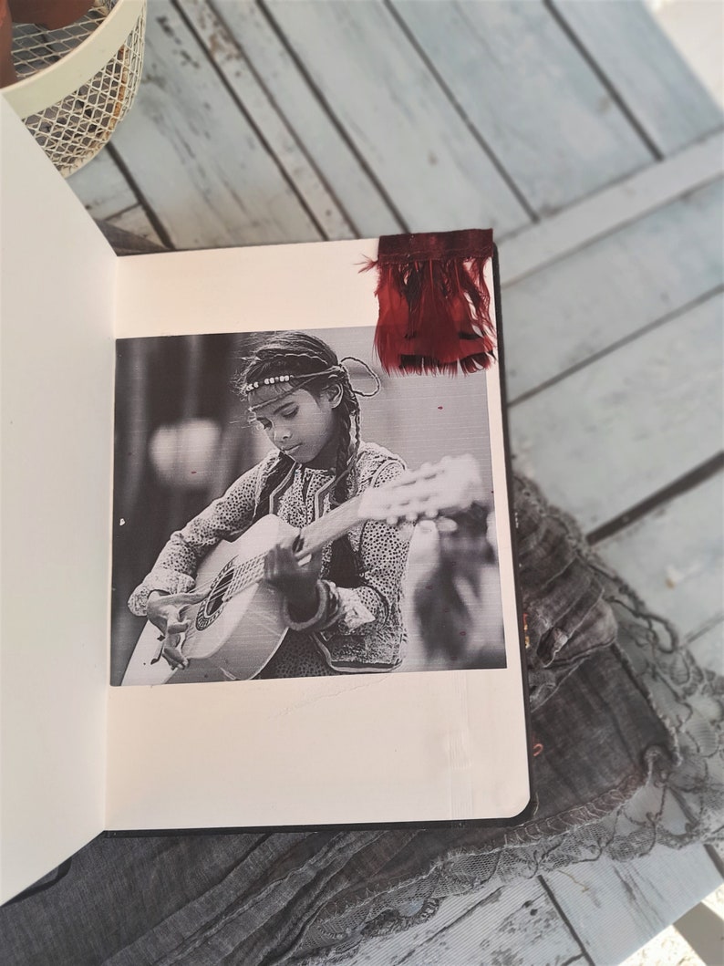 May include: A black and white photo of a person playing a guitar. The person is wearing a beaded headband and a patterned shirt. The photo is in a scrapbook with a red feather bookmark.