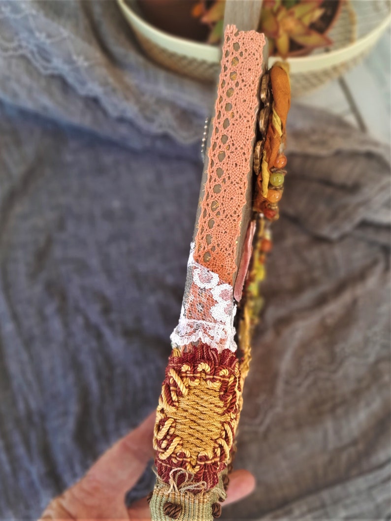 May include: A wooden frame with a decorative design featuring a peach-colored lace trim, white lace, and a gold and red woven trim. The frame is decorated with beads and other embellishments.