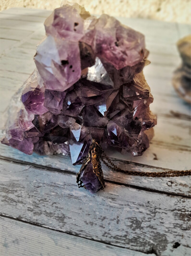 May include: A purple amethyst crystal pendant necklace with a detailed metal setting. The pendant is resting on a cluster of amethyst crystals.