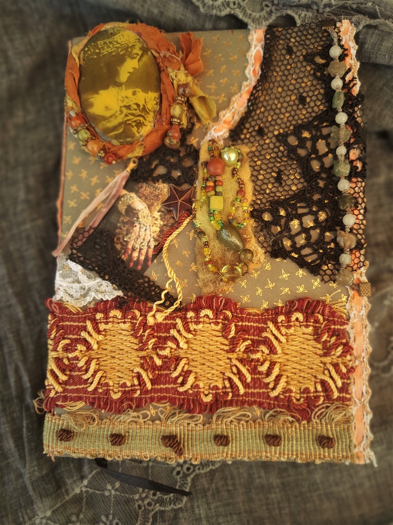May include: A handmade journal with a brown cover decorated with lace, fabric scraps, beads, and a gold-toned circular embellishment. The journal has a red and gold trim and a black ribbon.