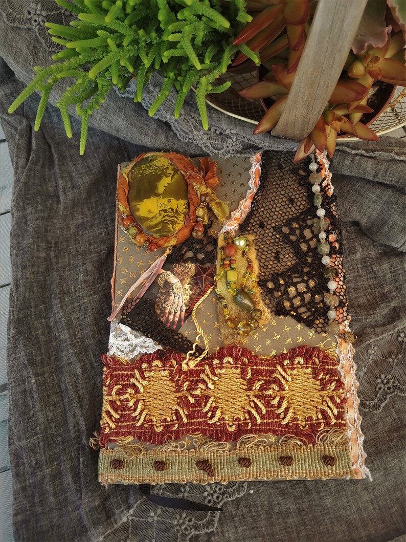 May include: A handmade journal with a brown cover decorated with lace, beads, and a gold-toned mirror. The journal has a rustic, vintage aesthetic.