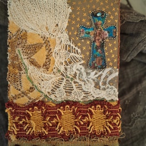 May include: A handmade journal cover with a cross design. The cross is made of fabric and has a blue and gold color scheme. The cover is decorated with lace and fabric scraps.