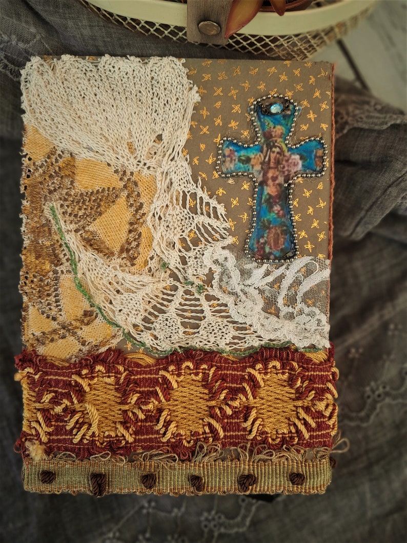 May include: A handmade journal cover with a cross design. The cross is made of fabric and has a blue and gold color scheme. The cover is decorated with lace and fabric scraps.