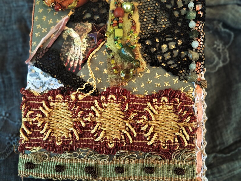 May include: A handmade journal cover with a variety of textures and embellishments, including lace, beads, and fabric. The cover is decorated with a star, a hand, and a beaded necklace.