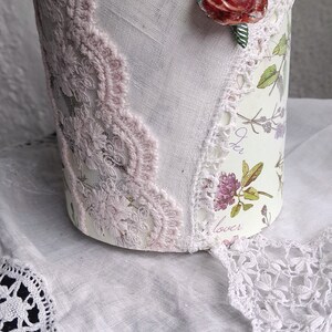Puede incluir: Un recipiente cilíndrico con un diseño floral, adornado con encaje rosa y un adorno de rosa roja. El recipiente está decorado con un estampado botánico y delicados adornos de encaje, creando una estética vintage.