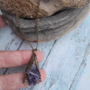 May include: A purple amethyst crystal pendant necklace with an ornate bronze filigree setting. The necklace is on a chain with a bronze finish.