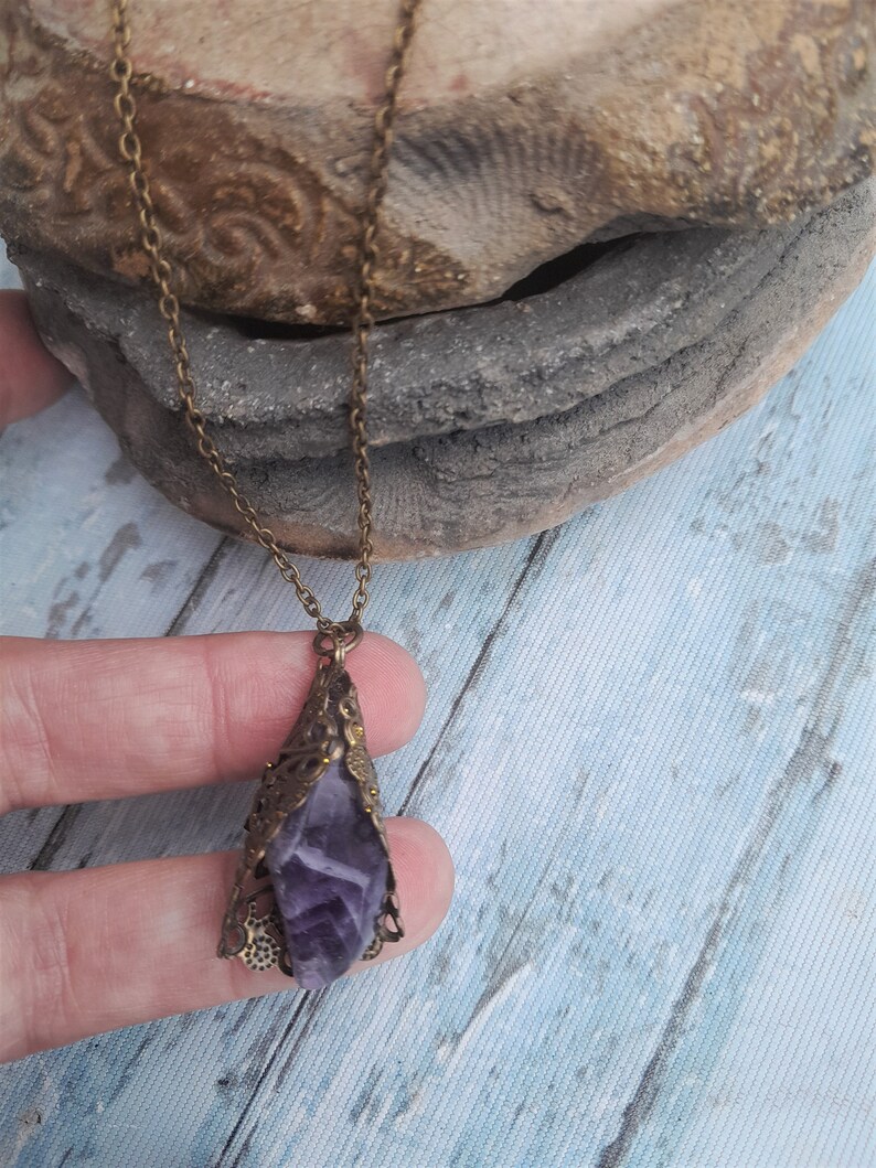 May include: A purple amethyst crystal pendant necklace with an ornate bronze filigree setting. The necklace is on a chain with a bronze finish.