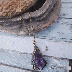 May include: A purple amethyst crystal pendant necklace with an ornate bronze filigree setting. The necklace is hanging from a bronze chain.