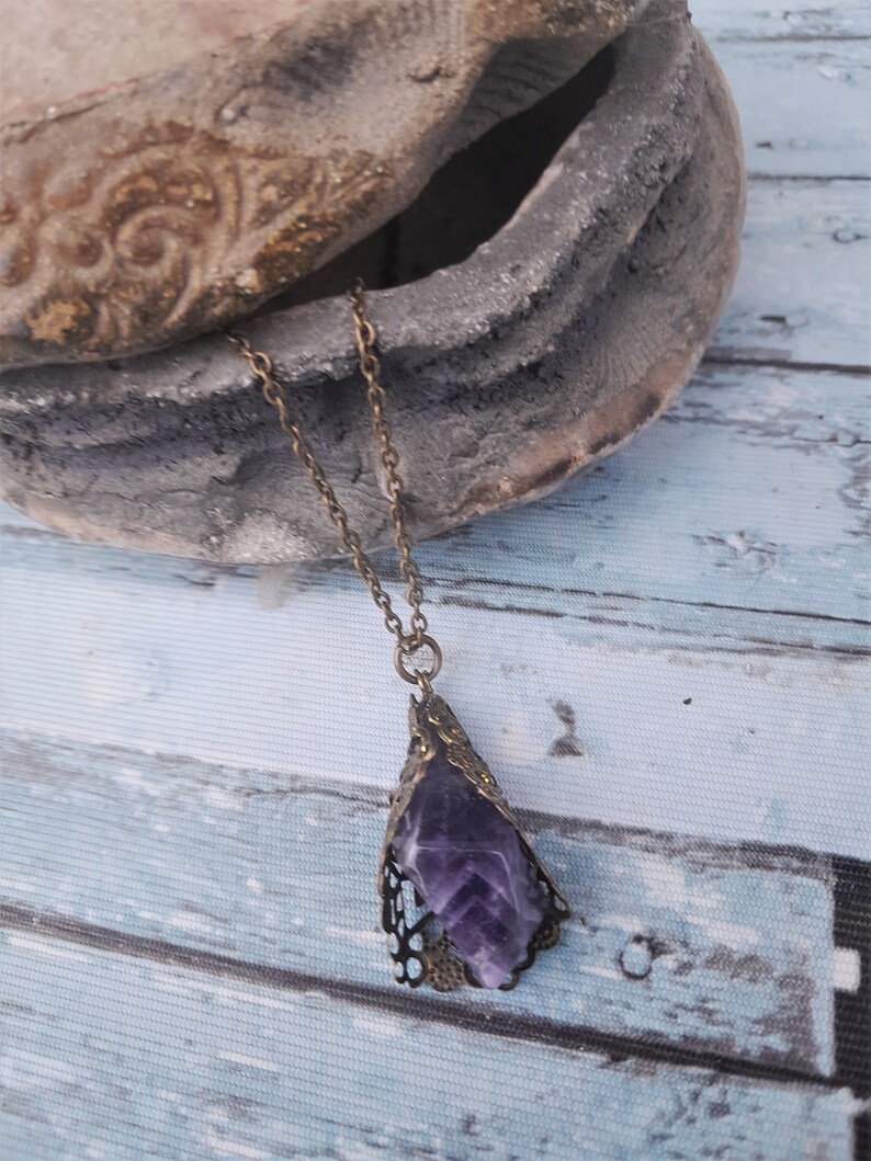 May include: A purple amethyst crystal pendant necklace with an ornate bronze filigree setting. The necklace is hanging from a bronze chain.