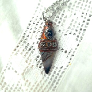 Peut inclure: Un collier pendentif avec une pierre d'agate en forme de cône et une chaîne en argent. Le pendentif a un boîtier en métal orange et argent avec une pierre bleu foncé et de petits cristaux décoratifs. Le collier est présenté sur un tissu blanc en forme de dentelle.