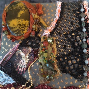 May include: A small, square, gray box with a gold star pattern. The box is decorated with a variety of materials, including lace, fabric, beads, and stones. The box has a vintage, eclectic aesthetic.