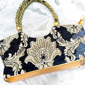 May include: A vintage handbag with a dark blue and gold floral pattern. The bag features a gold rope handle and a light brown leather base. The design is ornate and elegant, suitable for special occasions.