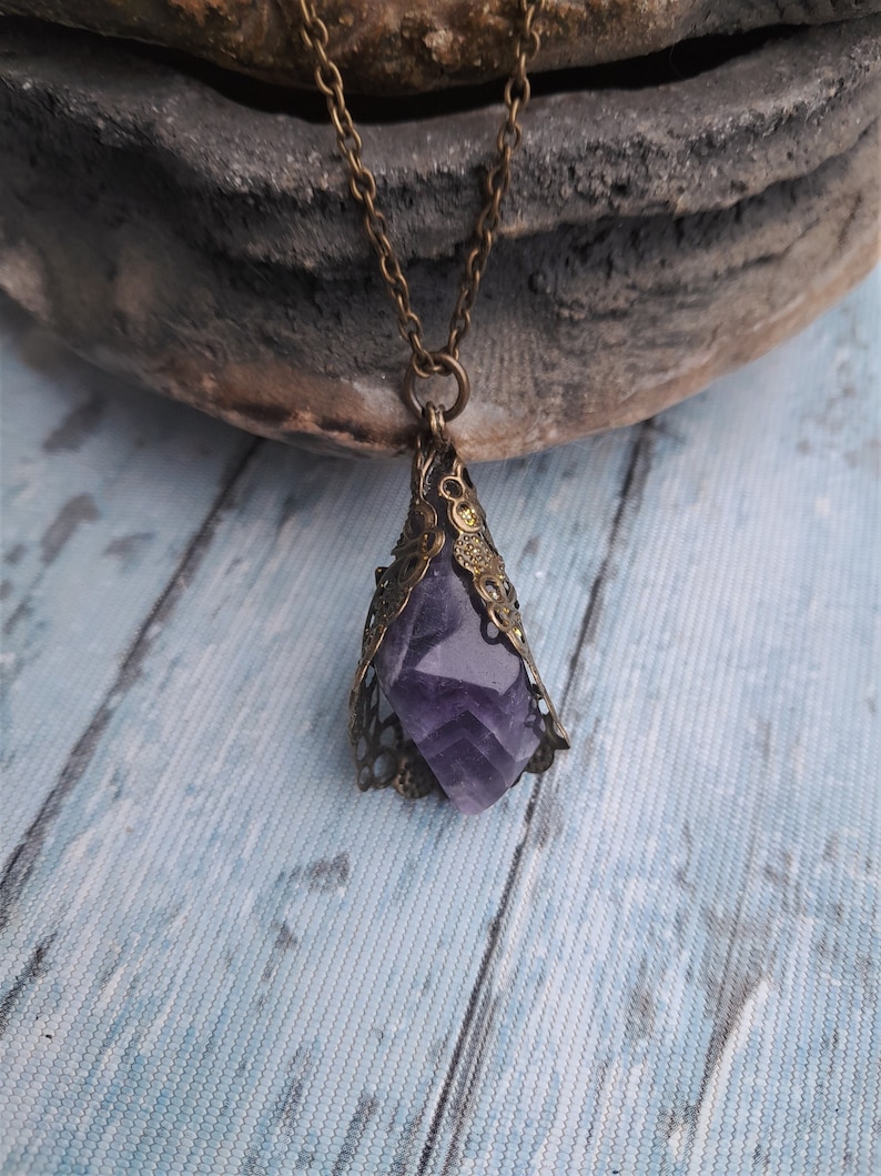 May include: A purple amethyst crystal pendant necklace with an ornate bronze filigree setting. The necklace is on a chain with a bronze finish.