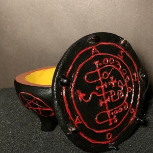 Puede incluir: Caja de cerámica negra con un sello rojo y negro en la tapa. La caja está abierta y el interior es amarillo.