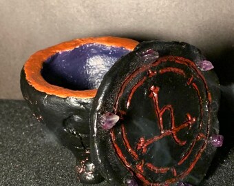 May include: Black ceramic incense burner with a lid featuring a red circle with a black symbol and purple crystals around the edge. The inside of the burner is purple.