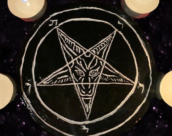 May include: A black, circular decorative item featuring a white pentagram with a goat-like face in the center. The pentagram is surrounded by a white border and symbols. Four lit, white candles are arranged around the item.