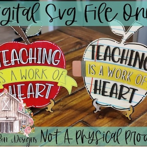 May include: Two red and white apple-shaped signs with the text "Teaching is a work of heart" on a wooden surface. The signs are decorated with a yellow ribbon and a pencil.