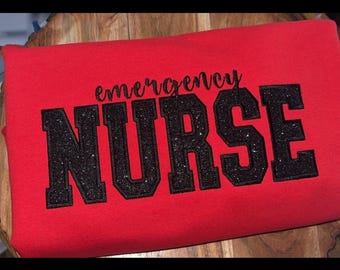 May include: A red sweatshirt with the words "emergency NURSE" in black glitter lettering. The text is in a bold, collegiate-style font. The sweatshirt is folded and appears to be made of a soft material.