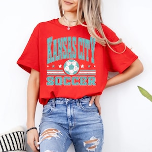 May include: A red t-shirt with the text "KANSAS CITY SOCCER" in turquoise. The design includes a soccer ball and stars. The shirt is being worn with blue jeans.