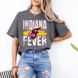 Puede incluir: Una camiseta gris carb&oacute;n de manga corta con un estampado gr&aacute;fico desgastado que dice INDIANA FEVER en letras blancas. Una letra F estilizada en azul marino y rojo incorpora un dise&ntilde;o de baloncesto sobre una barra amarilla.