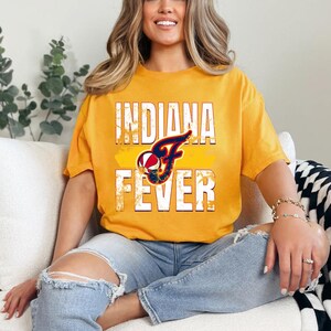 Puede incluir: Una camiseta de manga corta color amarillo mostaza con el texto INDIANA FEVER en letras grandes de bloque blancas desgastadas. Centrada entre las palabras hay una letra F azul marino estilizada con un gr&aacute;fico de baloncesto integrado.
