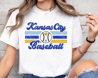 May include: White t-shirt with a blue and yellow graphic design that says "Kansas City Baseball" with a baseball and crown graphic.