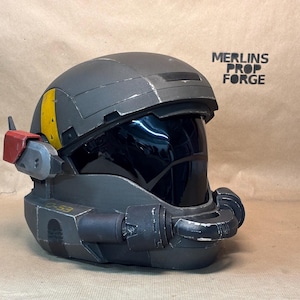 Op de afbeelding: Een donkergrijze tactische helm met een glanzend zwart vizier, een gele streep aan de zijkant en een rood accentstuk. De helm bevat een op de kin gemonteerd apparaat en de tekst "MERLINS PROP FORGE" en "C-53" op het buitenoppervlak.