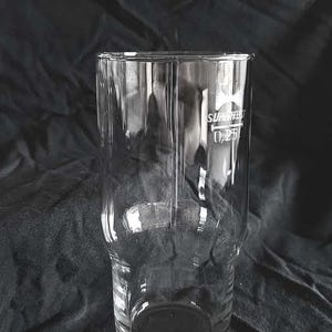 Könnte beinhalten: Ein klares, zylindrisches Bierglas mit breiterem Boden. Auf dem Glas befinden sich ein Logo und der Text "SUPERTEST 0,25l". Das leere Glas steht vor einem dunklen, strukturierten Hintergrund.