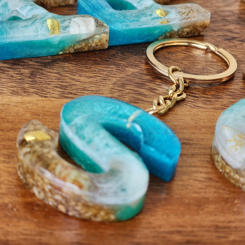 May include: A blue and white resin letter S keychain with gold flecks and a gold chain. The letter is made to look like ocean waves.