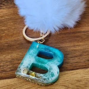 May include: A white fluffy keychain with a gold key ring attached to a blue and white resin letter B. The letter B has a beach scene inside with seashells and sand.