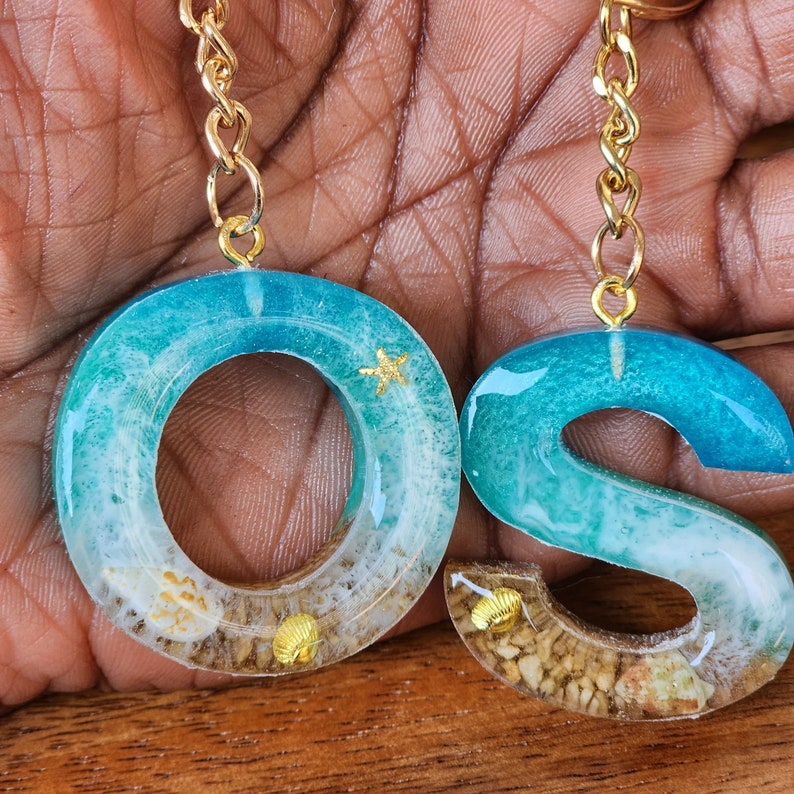 May include: A pair of resin keychains shaped like the letters "O" and "S". The keychains are blue and white with gold glitter and seashells embedded in the resin.
