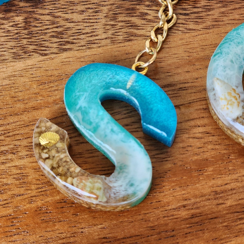 May include: A turquoise and white resin letter S keychain with a gold chain and seashells embedded in the resin.
