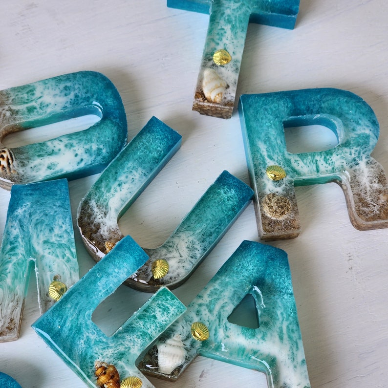 May include: A set of resin letters in a blue and white ocean wave design. The letters are decorated with gold accents and seashells.
