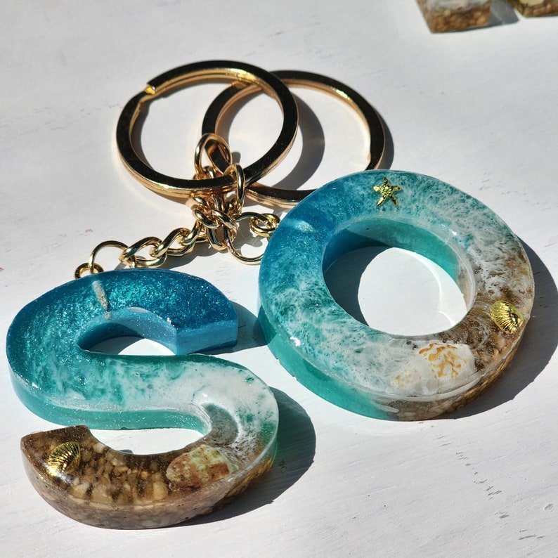 May include: A gold key chain with two blue and white resin charms shaped like the letters "S" and "O". The charms have a beachy theme with seashells and starfish.