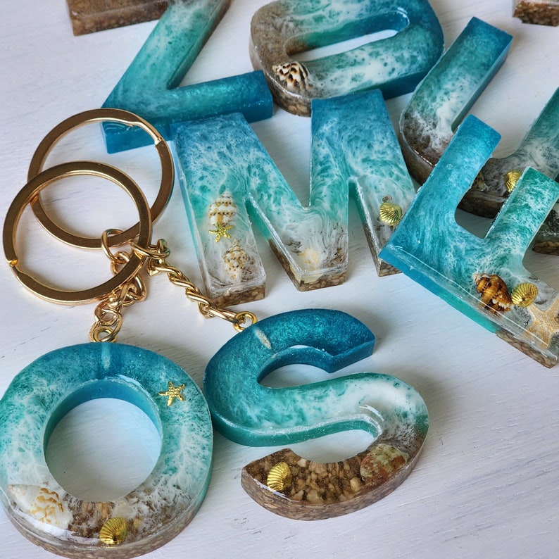 May include: A turquoise and white resin keychain with a gold key ring. The keychain is shaped like the letters "O" and "S" and features a beachy design with seashells and a wave pattern.