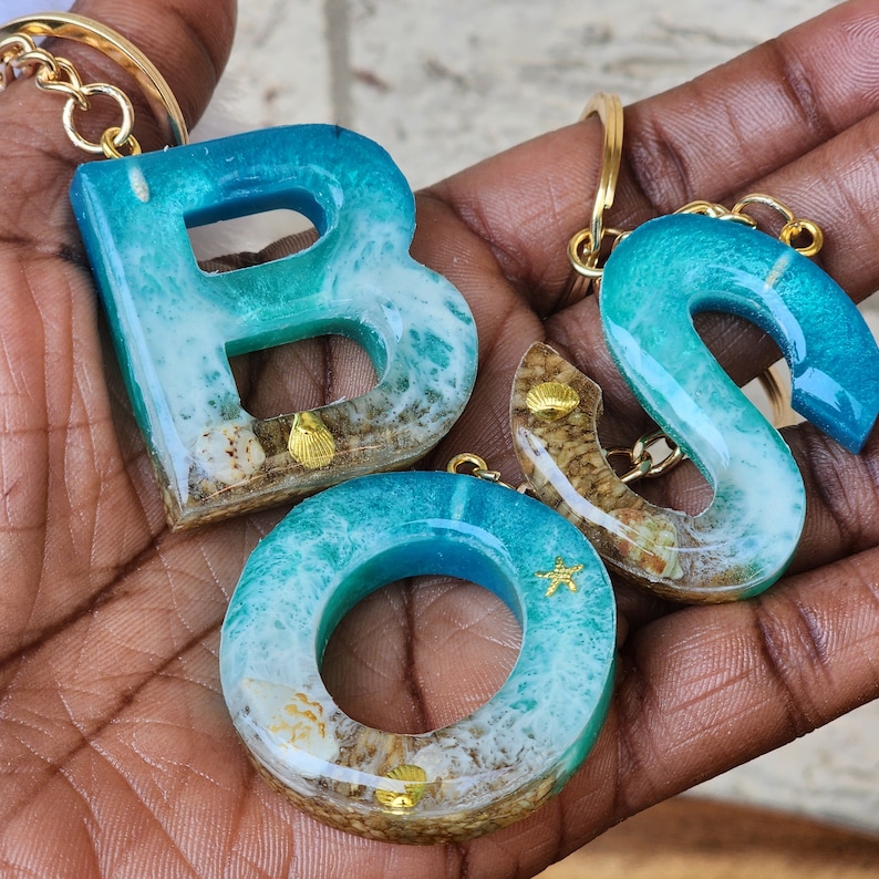 May include: Three resin keychains shaped like the letters B, O, and S. The keychains are blue and white with gold accents and seashells embedded in the resin.