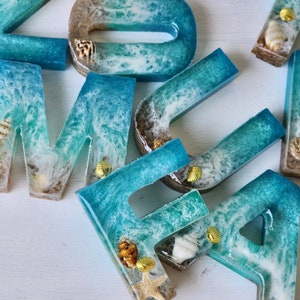 May include: A set of resin letters in the shape of the word "HOME" with a blue and white ocean wave design. The letters are decorated with seashells and gold accents.