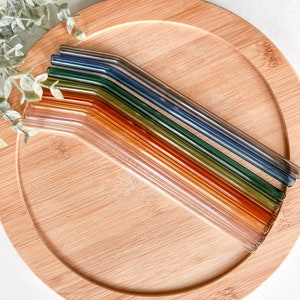 May include: A collection of colorful glass straws in shades of orange, green, and blue, arranged on a round bamboo tray. The straws are angled and appear to be reusable. The tray is light brown and has a natural wood grain.