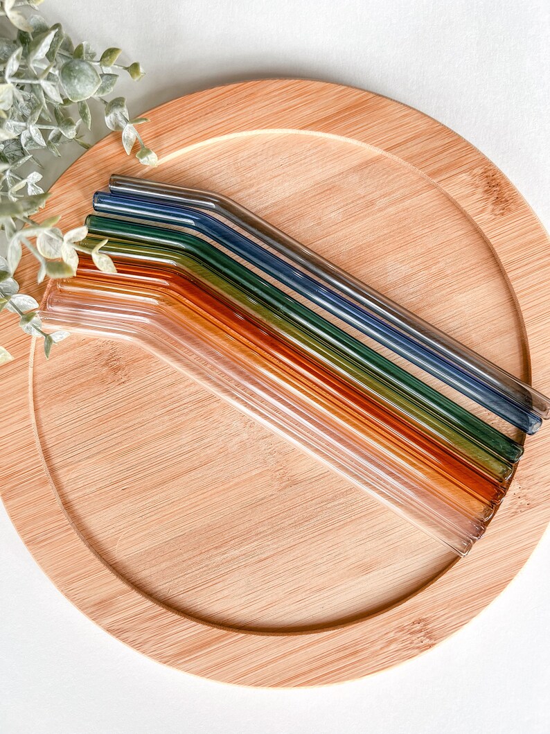 May include: A collection of colorful glass straws in shades of orange, green, and blue, arranged on a round bamboo tray. The straws are angled and appear to be reusable. The tray is light brown and has a natural wood grain.