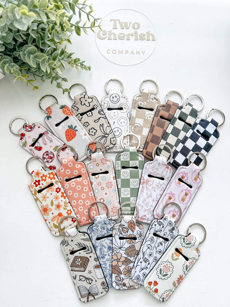 K&ouml;nnte beinhalten: Eine Sammlung von Lippenbalsamhaltern mit Schl&uuml;sselringen und verschiedenen Mustern. Designs umfassen Kirschen, Erdbeeren, Smiley-Gesichter, Blumenmuster und Schachbrettmuster. Die Halter sind in verschiedenen Farben erh&auml;ltlich, darunter Rosa, Wei&szlig; und Beige. Der Markenname "Two Cherish Company" ist sichtbar.