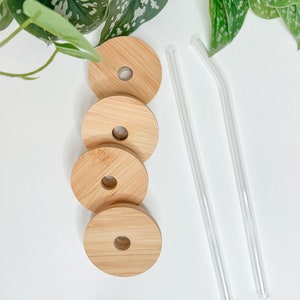May include: Four round bamboo lids with a hole in the center and two clear glass straws. The lids are light brown and have a natural wood grain. The straws are straight and curved. The items are arranged on a white surface.