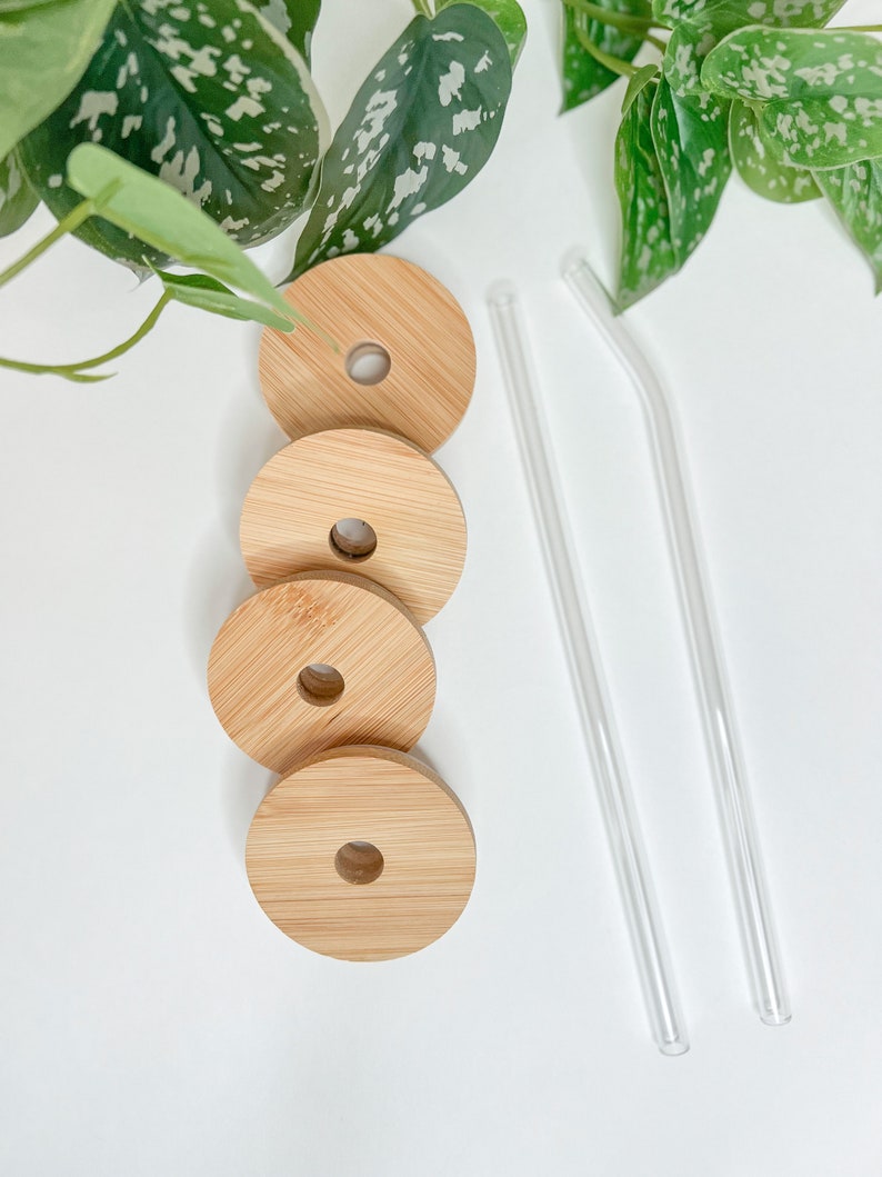 May include: Four round bamboo lids with a hole in the center and two clear glass straws. The lids are light brown and have a natural wood grain. The straws are straight and curved. The items are arranged on a white surface.