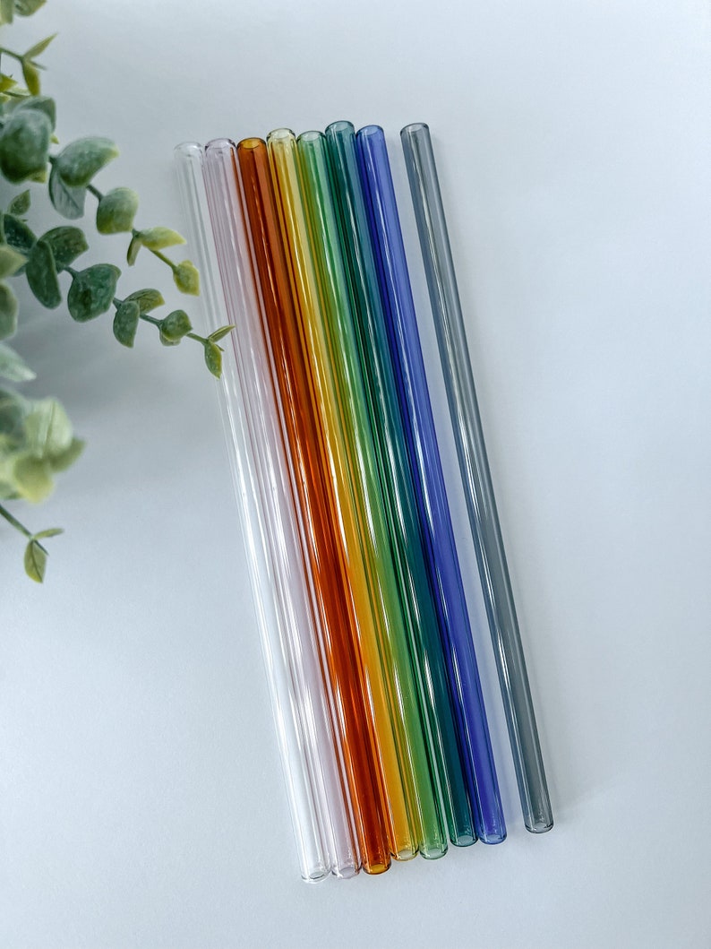 May include: A collection of ten colorful glass straws arranged diagonally on a white surface. The straws are clear, with hues of pink, orange, yellow, green, blue, and gray. A green leafy plant is visible in the upper left corner.