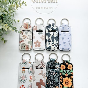 K&ouml;nnte beinhalten: Acht Schl&uuml;sselanh&auml;nger mit verschiedenen Mustern, darunter B&uuml;cher, Blumen, Schleifen und Sonnenblumen. Jeder Schl&uuml;sselanh&auml;nger hat einen silbernen Ring und einen wei&szlig;en Hintergrund. Der Markenname "Two Cherish Company" ist sichtbar.