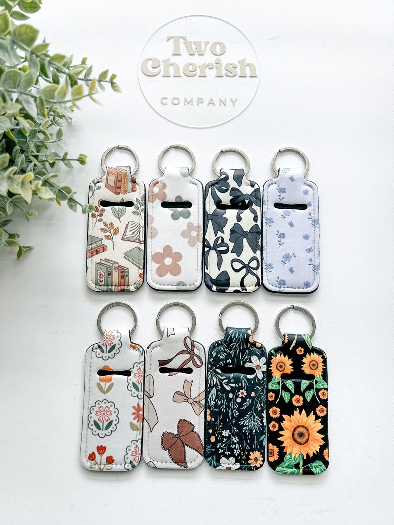 K&ouml;nnte beinhalten: Acht Schl&uuml;sselanh&auml;nger mit verschiedenen Mustern, darunter B&uuml;cher, Blumen, Schleifen und Sonnenblumen. Jeder Schl&uuml;sselanh&auml;nger hat einen silbernen Ring und einen wei&szlig;en Hintergrund. Der Markenname "Two Cherish Company" ist sichtbar.
