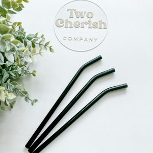 May include: Three dark green, curved metal straws are arranged diagonally on a white surface. The straws are slightly bent near the top. A clear circular sign with the text "Two Cherish Company" is in the background.