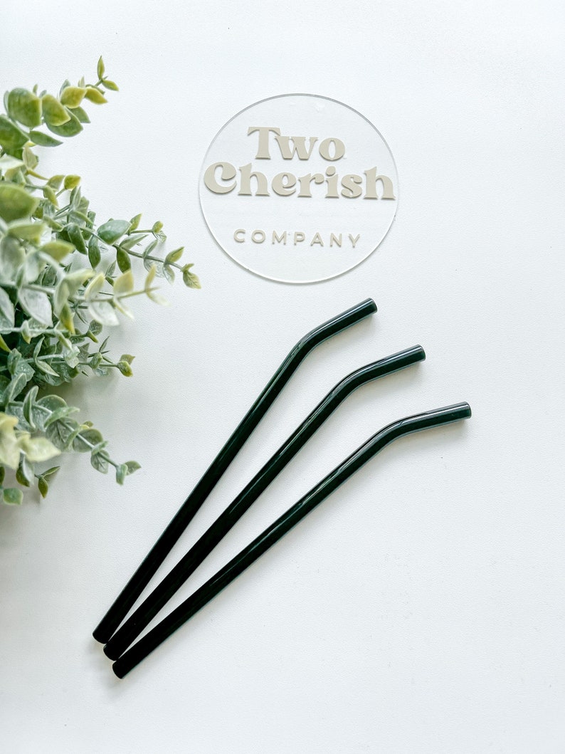 May include: Three dark green, curved metal straws are arranged diagonally on a white surface. The straws are slightly bent near the top. A clear circular sign with the text "Two Cherish Company" is in the background.