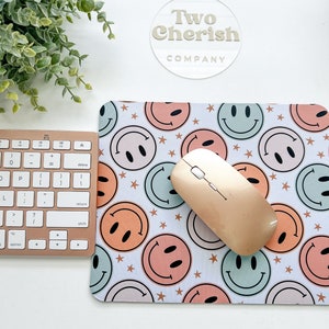 May include: A mousepad featuring a repeating pattern of pastel-coloured smiley faces and stars. A wireless mouse and a rose gold keyboard sit on a white surface. The Two Cherish Company logo is visible in the background.