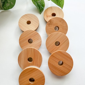 May include: A collection of eight round bamboo lids with a central hole, arranged on a white surface. The lids are a light brown color, showcasing the natural wood grain. Green foliage is visible in the background.